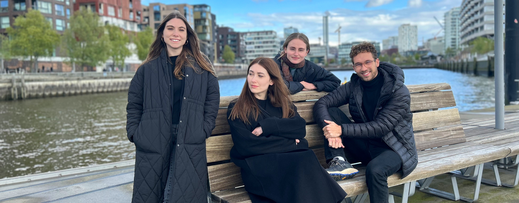 Die Gründerinnen und der Gründer von moneten sitzen auf einer Bank am Wasser.